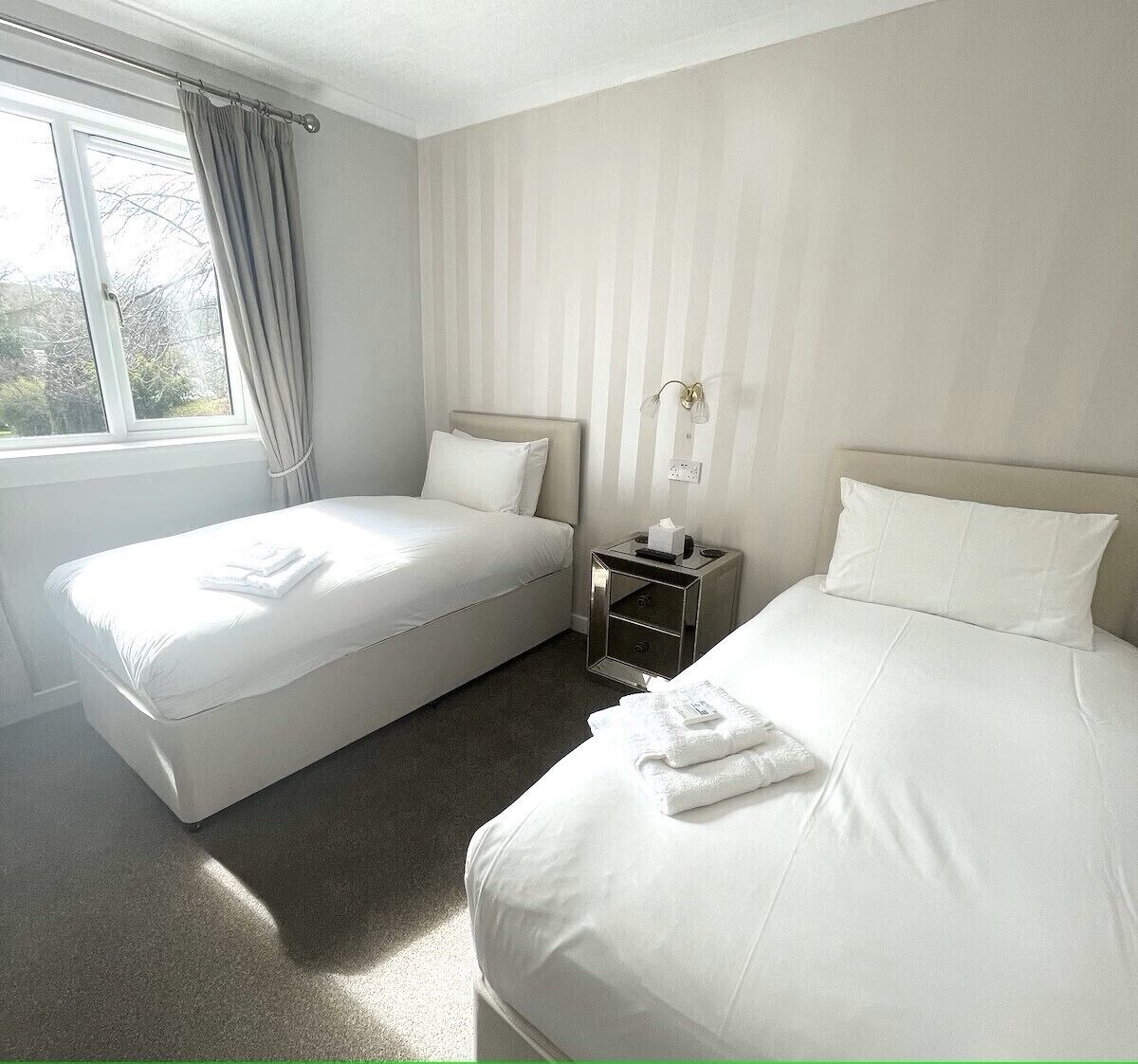 A hotel room with two beds, a nightstand, a window with curtains, and a wall-mounted light fixture.
