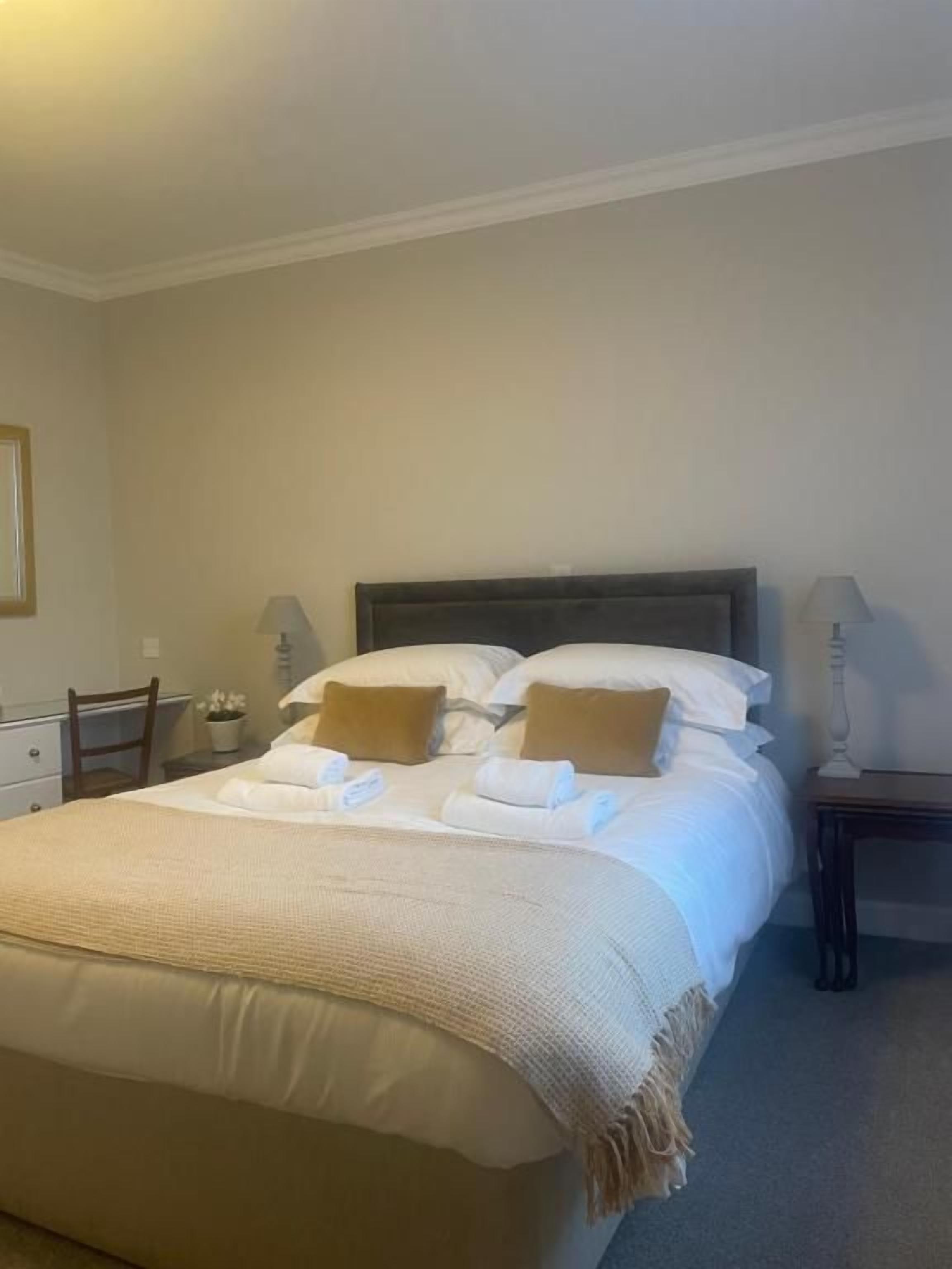 Ein ordentlich gemachtes Bett mit Kissen und Decke, ein Nachttisch mit Lampe sowie ein Sessel in einem Raum mit schlichten Wänden.