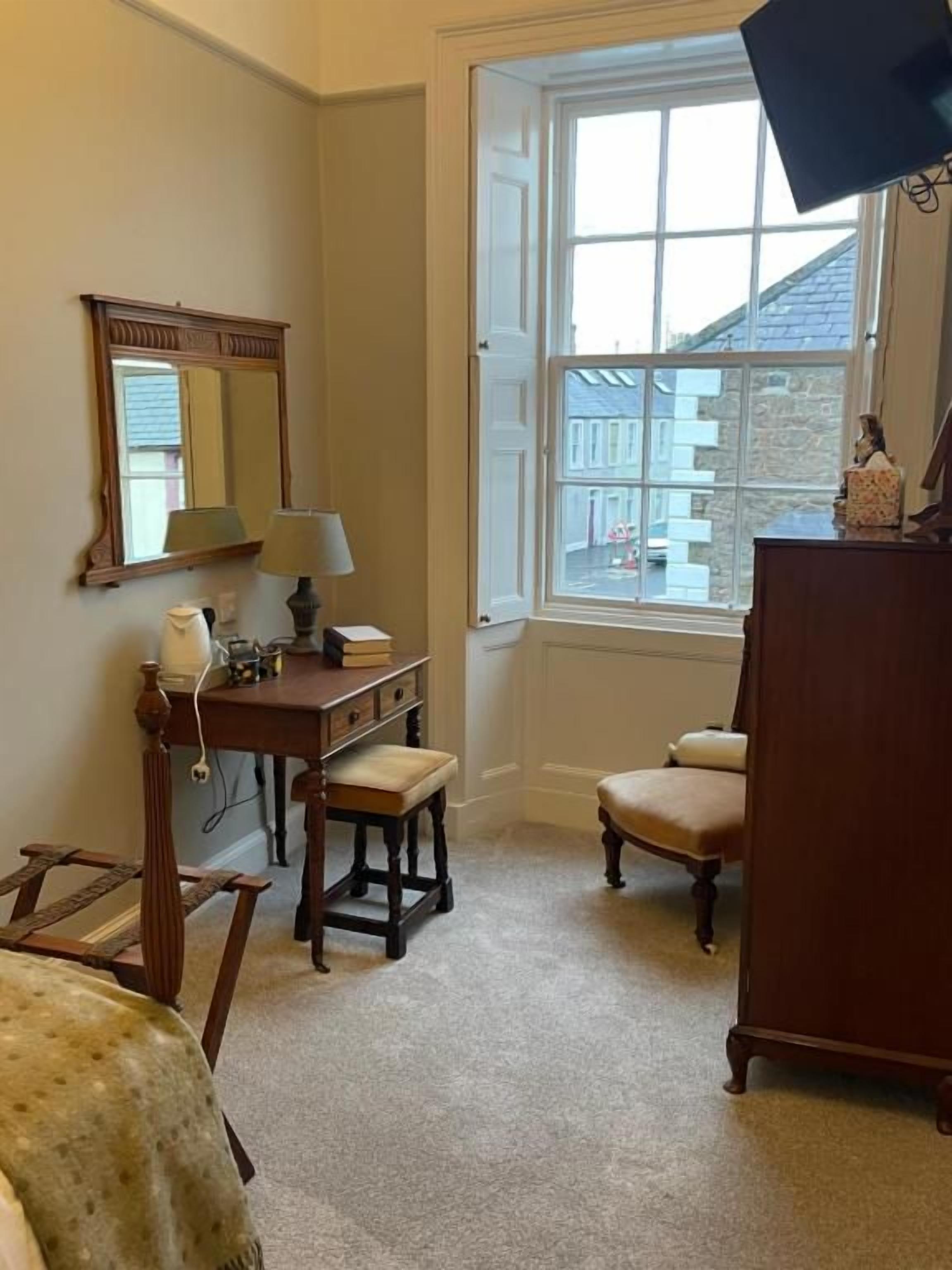 Une pièce comprenant un bureau en bois, une chaise, une petite table avec une lampe, un miroir et une fenêtre donnant sur des bâtiments.