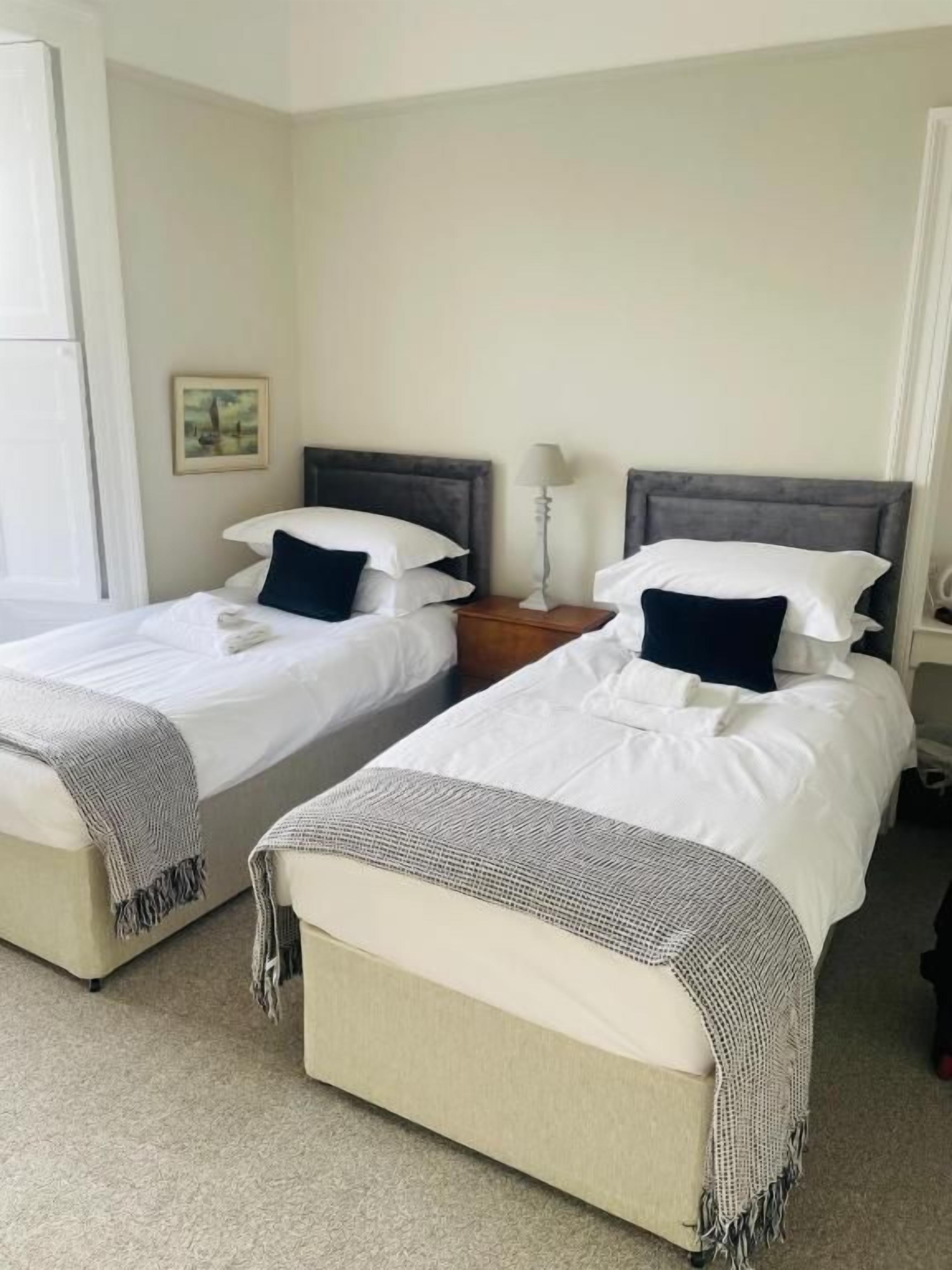 Habitación de hotel con dos camas, cada una con almohada y manta. Se observa una mesita de noche con lámpara entre las camas.