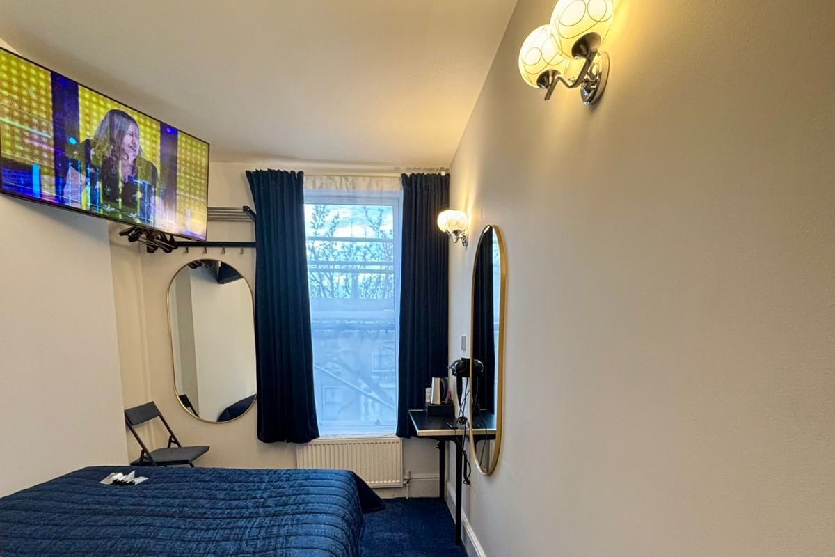 A hotel room with a bed, a TV, a mirror, a desk, and a window with curtains.