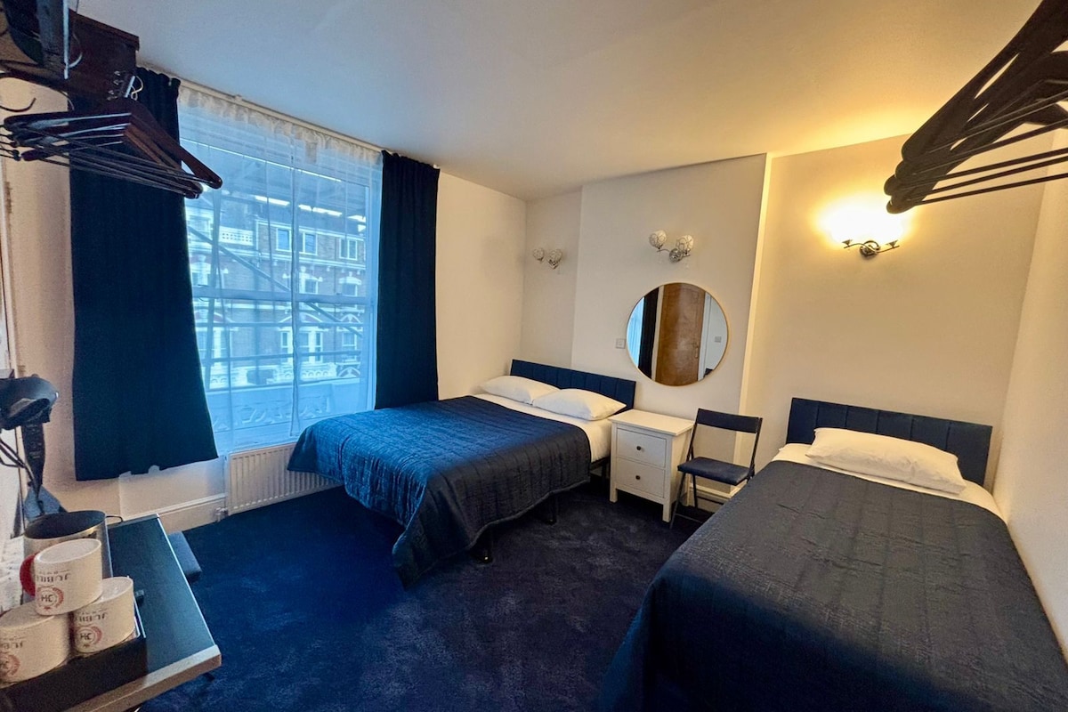 A hotel room with two beds, a small table, a chair, a mirror, and a window with curtains.