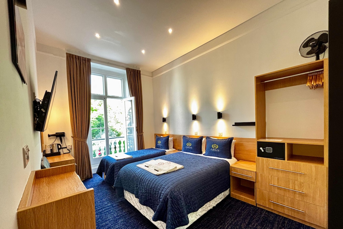 A hotel room with two beds, a desk, a safe, and a view of the outdoors.