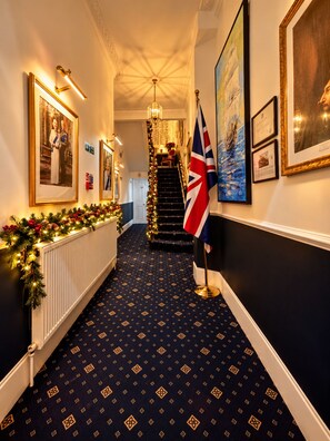 A hallway with a patterned carpet, framed pictures on the walls, a flag, and a staircase with a Christmas decoration. - Jubilee Hotel Victoria (London)