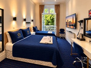 A hotel room with two beds, a desk, a chair, a television, and a window with a view of greenery. - Jubilee Hotel Victoria (London)