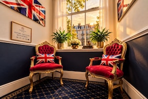 Two ornate chairs with Union Jack cushions, a framed sign indicating room numbers, and a window with a view of buildings. - Jubilee Hotel Victoria (London)
