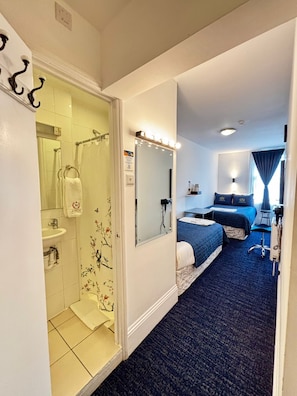 A hotel room with a shower, a bed with a blue bedspread, a desk with a laptop, and a mirror with lights above it. - Jubilee Hotel Victoria (London)