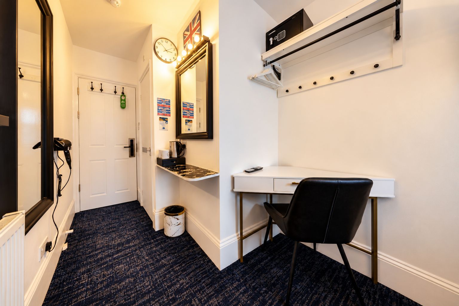 A hotel room with a white desk, a black chair, a clock, and a shelf with a kettle and a phone.