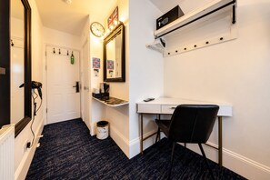 Una habitación de hotel con un escritorio blanco, una silla negra, un reloj y una estantería con una tetera y un teléfono.