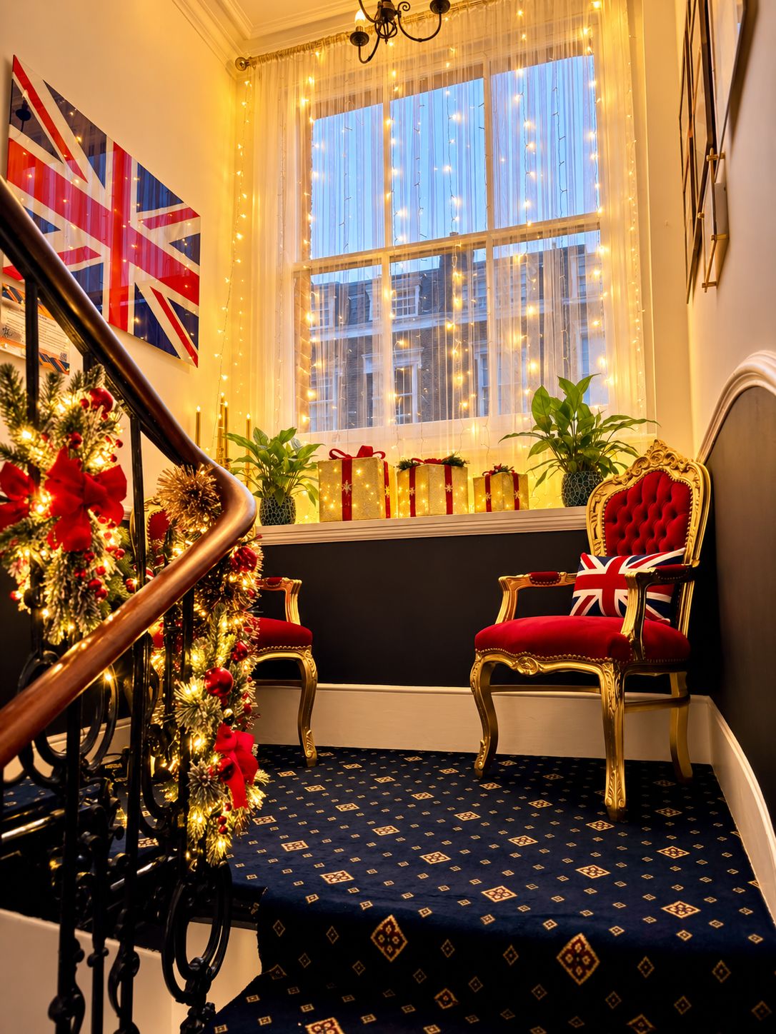 A staircase with a dark blue carpet, two ornate chairs with red upholstery, a window with string lights, and a British flag on the wall.