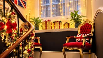 A staircase with a dark blue carpet, two ornate chairs with red upholstery, a window with string lights, and a British flag on the wall.
