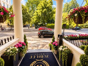 A hotel entrance with a black carpet and gold lettering, surrounded by greenery and a cobblestone path. - Jubilee Hotel Victoria (London)