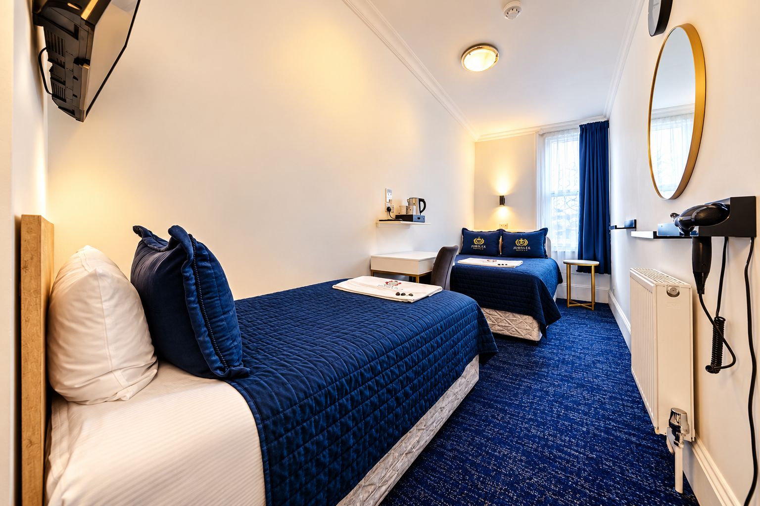 A hotel room with two beds, a desk, a mirror, and a radiator.