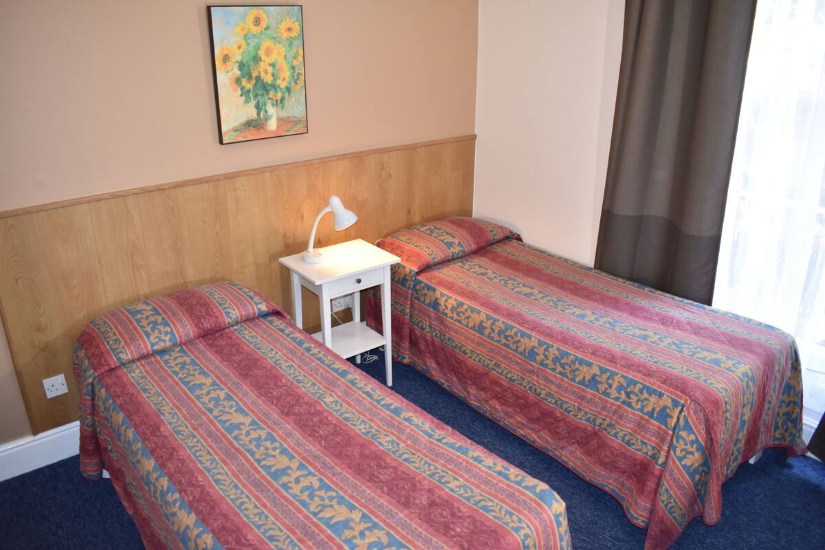A hotel room with two single beds, a nightstand with a lamp, a painting on the wall, and a window with curtains.