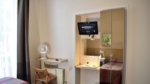 Habitación de hotel con cama, una silla, un escritorio con monitor de computadora, lámpara de escritorio, una mesita con un jarrón de flores y un armario con espejo.