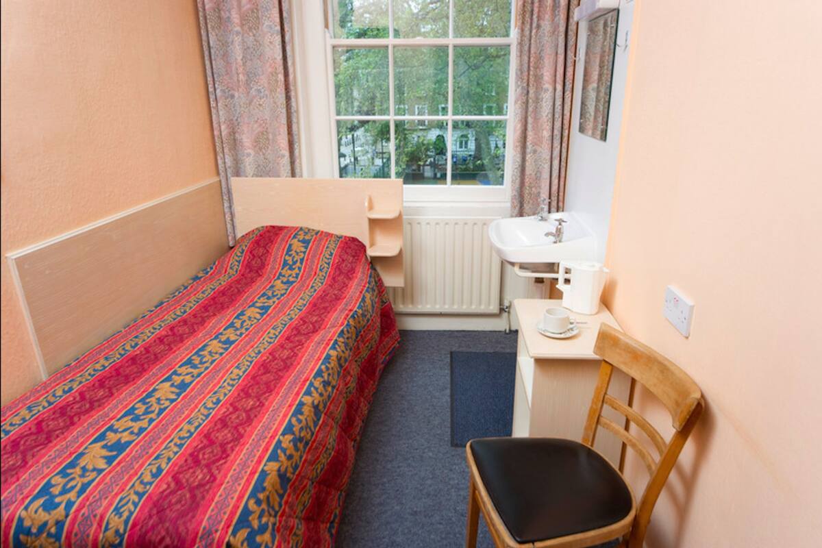 A small, neatly arranged room with a single bed, a wooden chair, a small table with a cup and saucer, and a window with curtains.