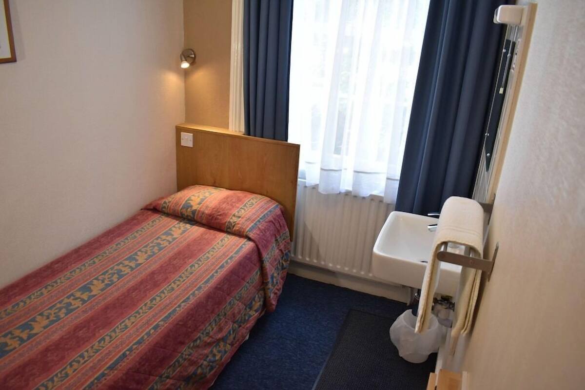 A small, single-bed room with a wooden headboard, a window with curtains, a white sink, and a towel rack.