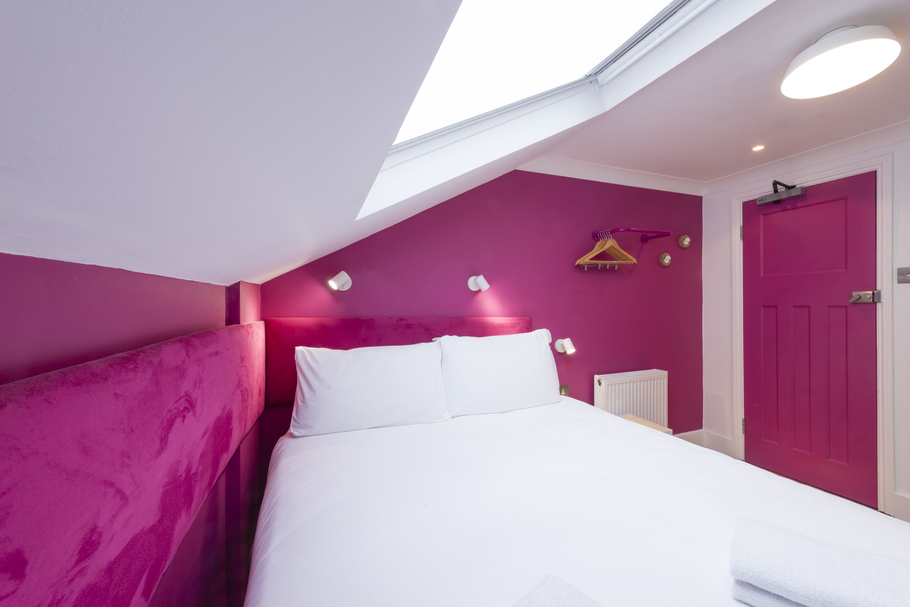 A bedroom with a skylight, a bed with white linens, a pink wall, and a door.