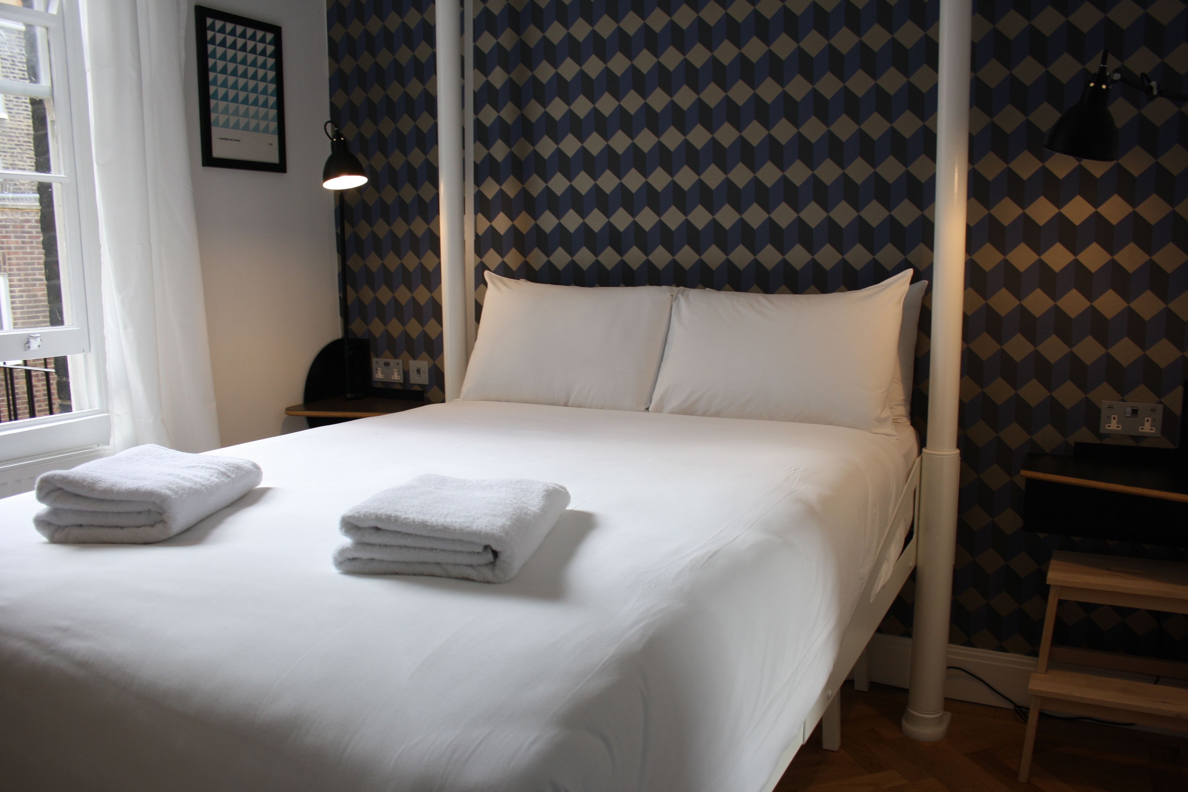 Una cama bien hecha con sábanas blancas y dos toallas dobladas, una lámpara de noche y un papel tapiz estampado.