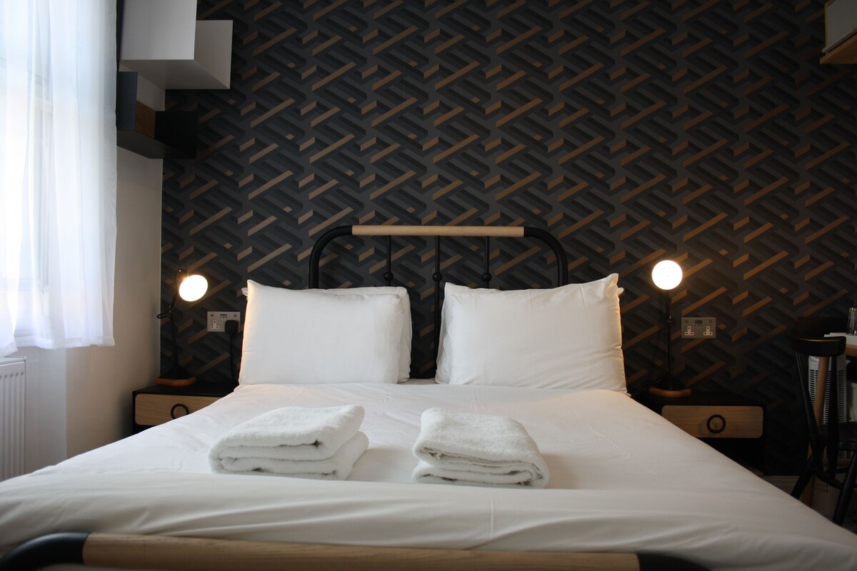 A neatly made bed with white linens and two folded towels, a wooden headboard, and a patterned wall.