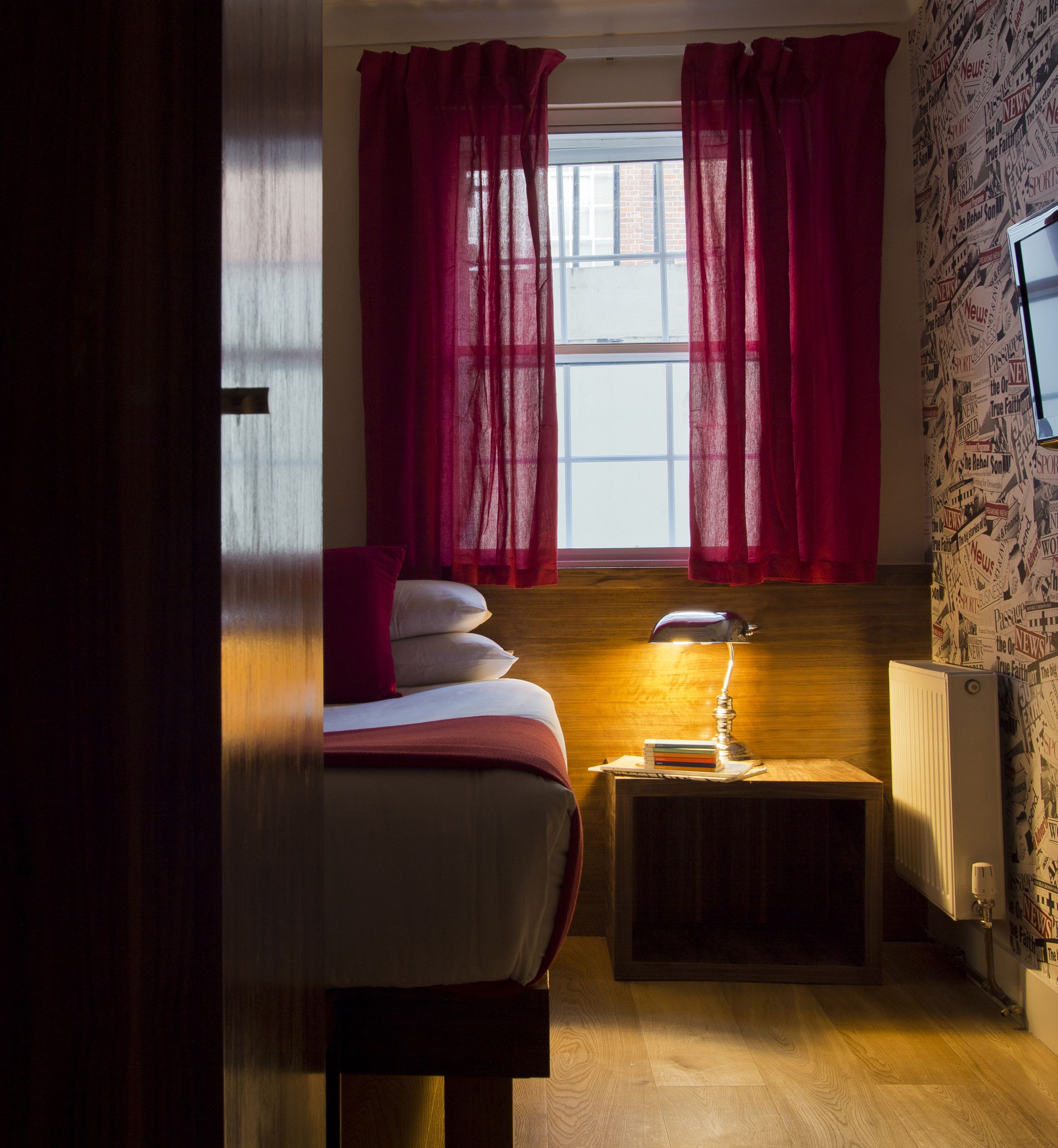 A bedroom with a bed, a desk with a lamp, a radiator, and a window with curtains.