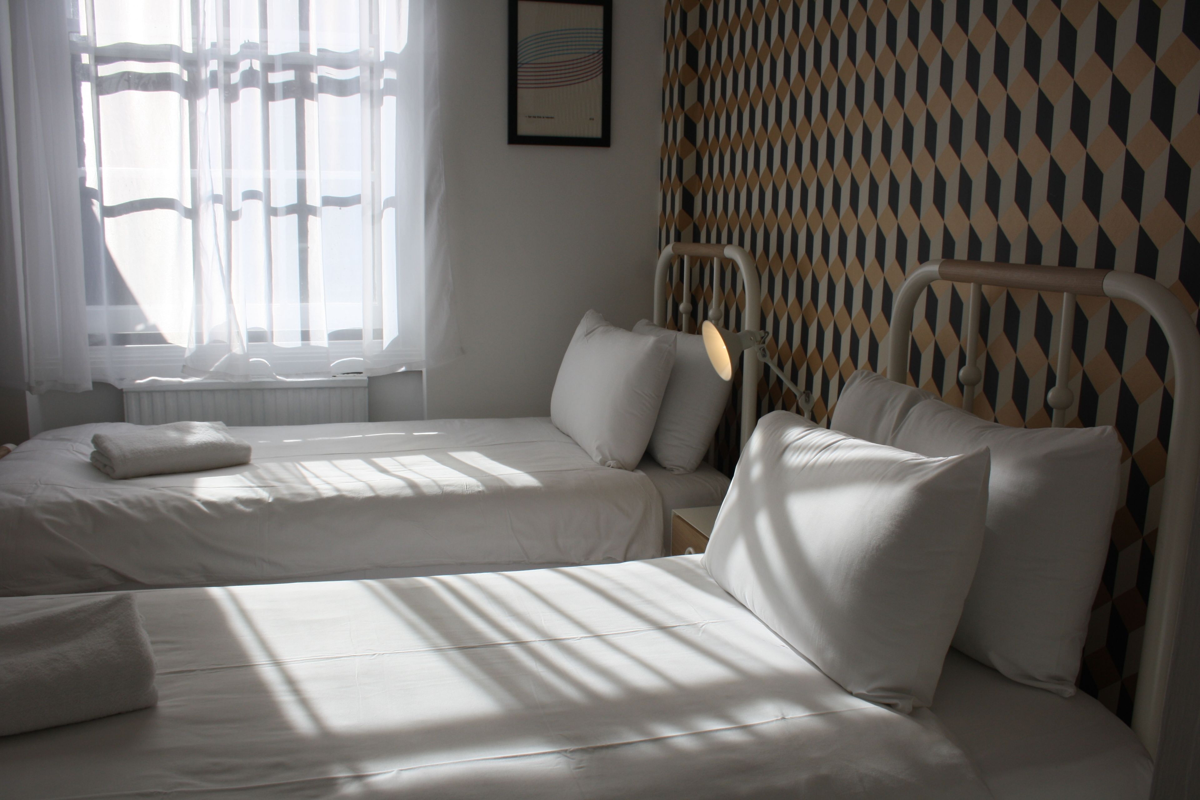 Une chambre d’hôtel avec deux lits, une fenêtre avec des rideaux légers, un mur à motifs et un tableau encadré accroché au mur.