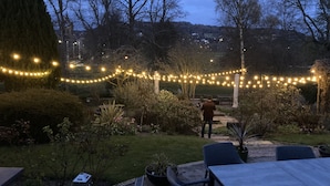 Un espace extérieur aménagé en terrasse, agrémenté de guirlandes lumineuses, d’une pelouse verdoyante et offrant une vue sur une colline en arrière-plan.