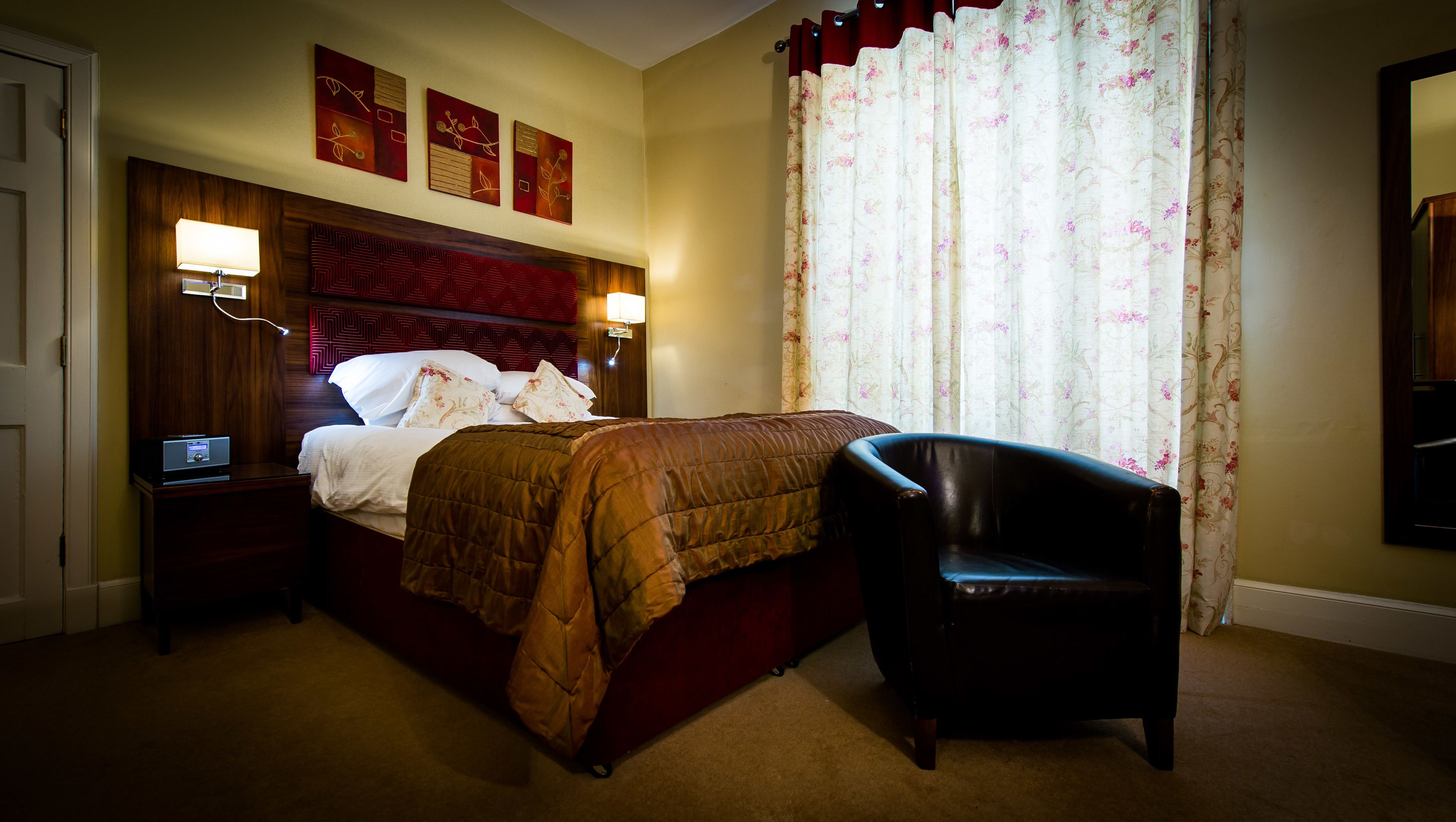 Une chambre d’hôtel avec un grand lit, une table de chevet avec une radio, une chaise et une fenêtre ornée de rideaux à motifs floraux.