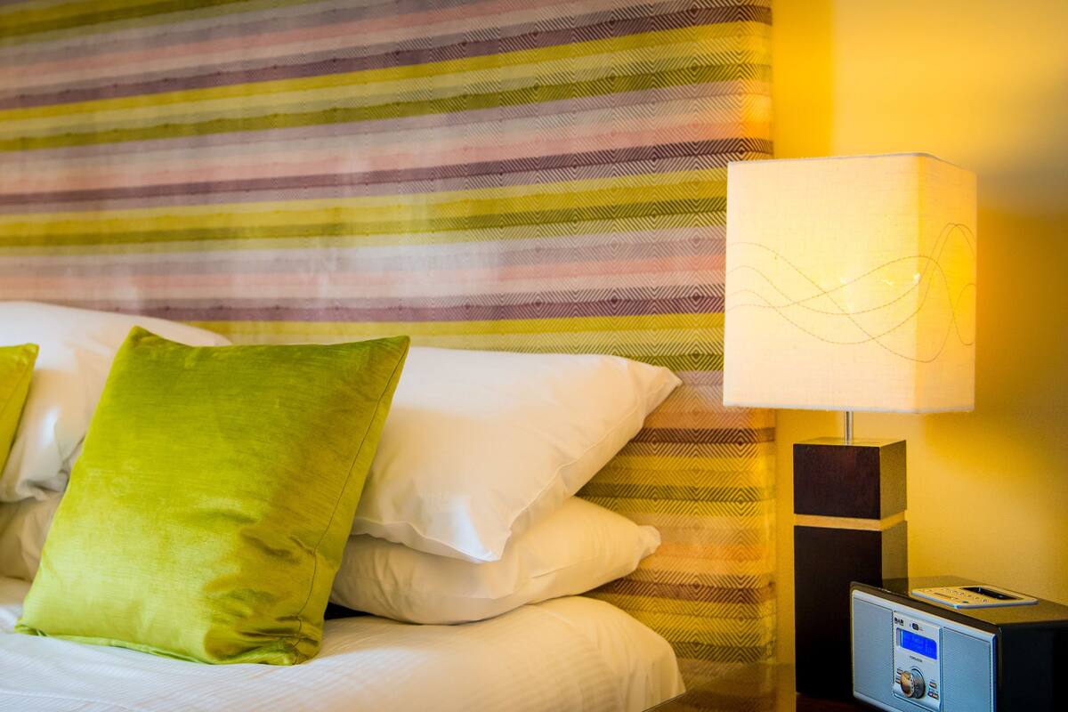 A neatly made bed with green and white pillows, a striped wall behind, and two bedside lamps.