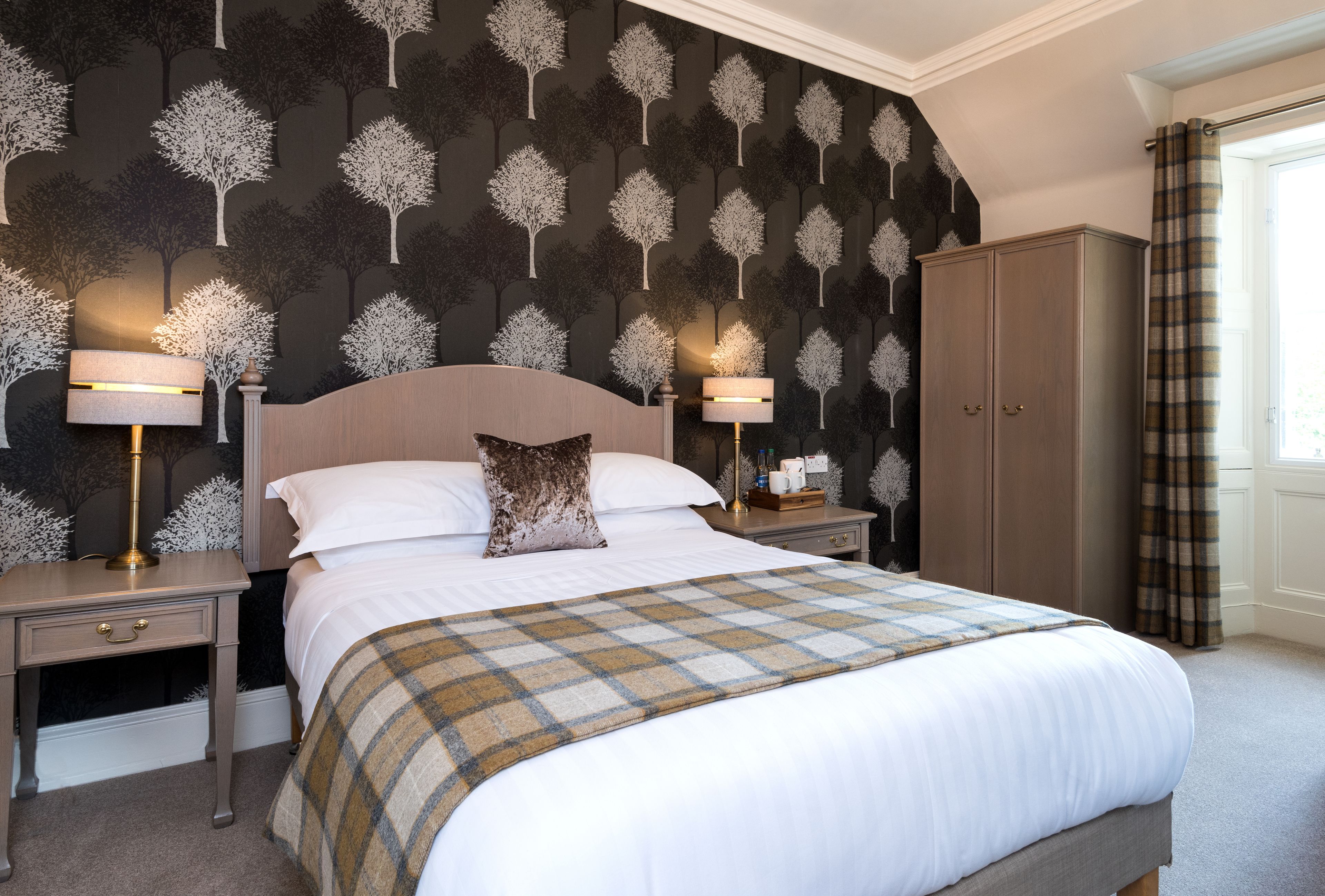 A neatly made bed with a plaid blanket, two bedside lamps, a nightstand, a wardrobe, and a patterned wallpaper.