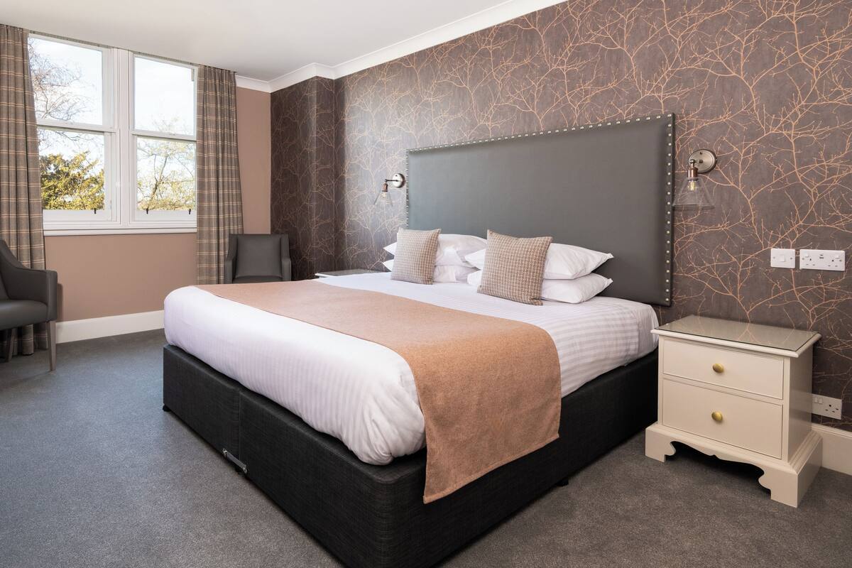 A modern hotel room with a large bed, a desk with a coffee maker, a chair, and a nightstand with a lamp.