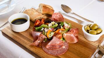 A wooden board with a variety of food items including sliced salami, prosciutto, a salad with mixed greens and vegetables, a bowl of olives, and a small bowl of olive oil.