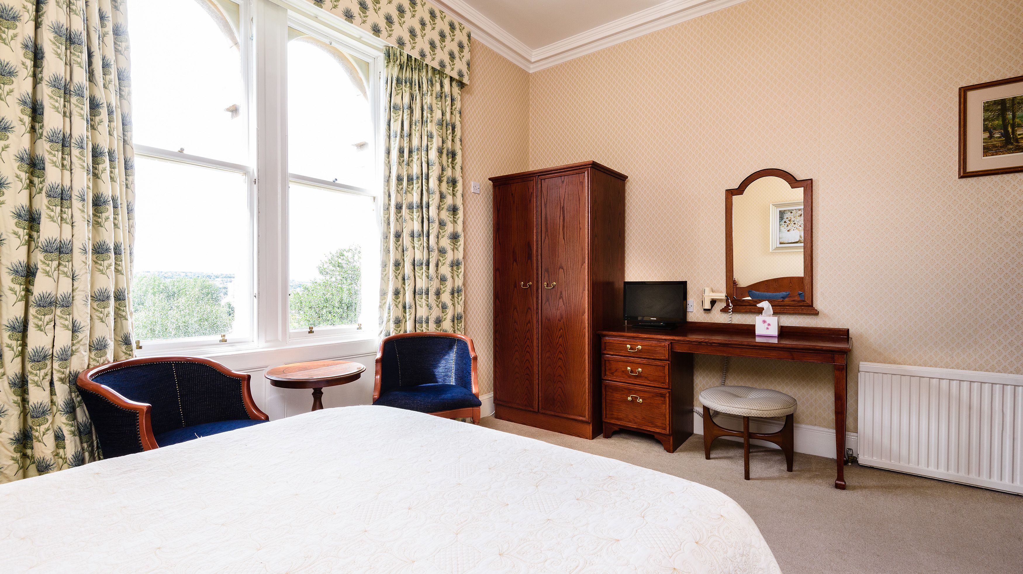 A hotel room with a large bed, a wooden desk, a chair, a small table, and a wardrobe.