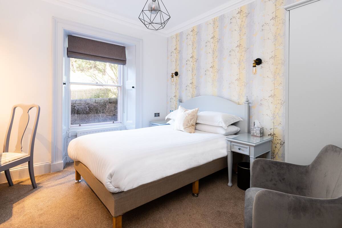A bedroom with a bed, a chair, a nightstand, and a window with a view of the outside.