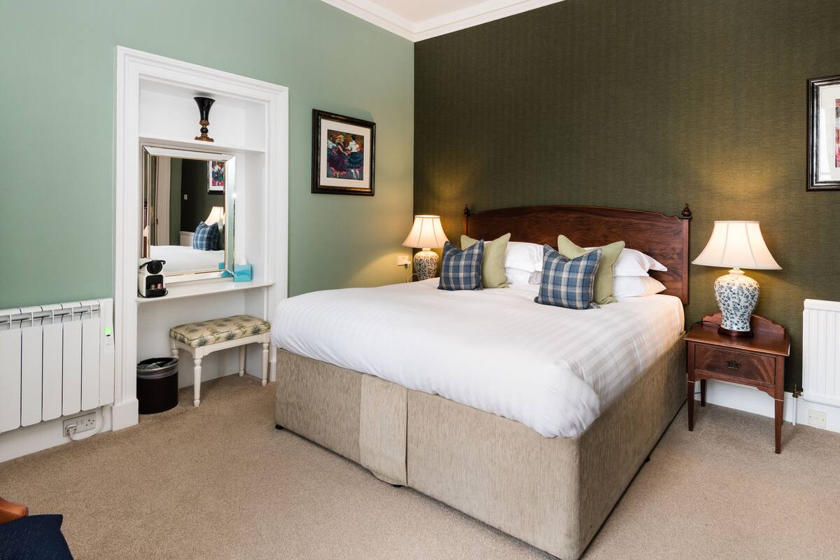 A neatly arranged bedroom with a large bed, bedside lamps, a dresser, a mirror, and a radiator.
