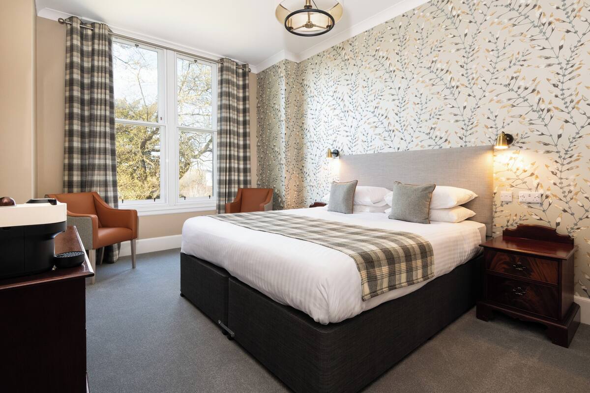 A bedroom with a large bed, a desk with a computer, a chair, a nightstand, and a window with curtains.