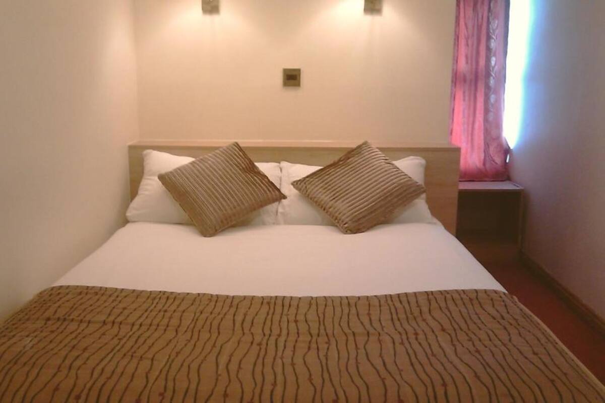 A neatly made bed with a striped blanket and two pillows, a bedside table with a lamp, and a window with curtains.