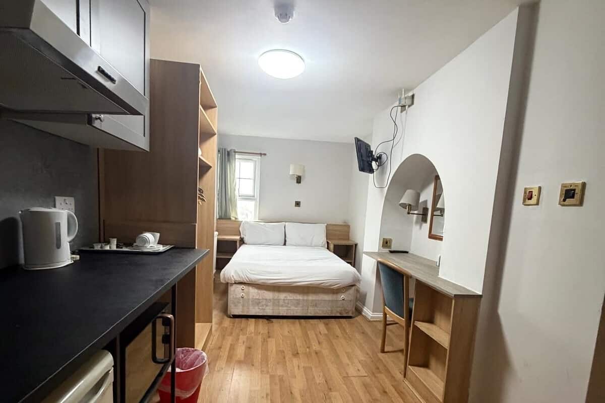 A compact room with a kitchenette, a bed, a desk, and a mirror.