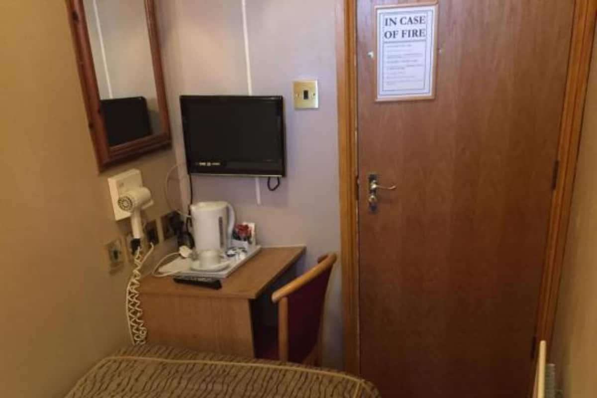 A hotel room with a bed, a desk with a computer monitor, a telephone, and a coffee maker. There is a door with a sign that reads "IN CASE OF FIRE".