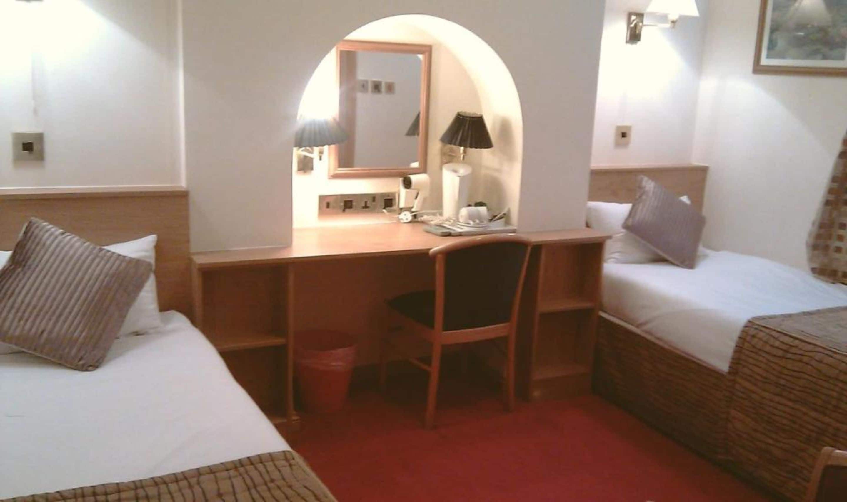Una camera d'albergo con una scrivania, due letti, una sedia e uno specchio.