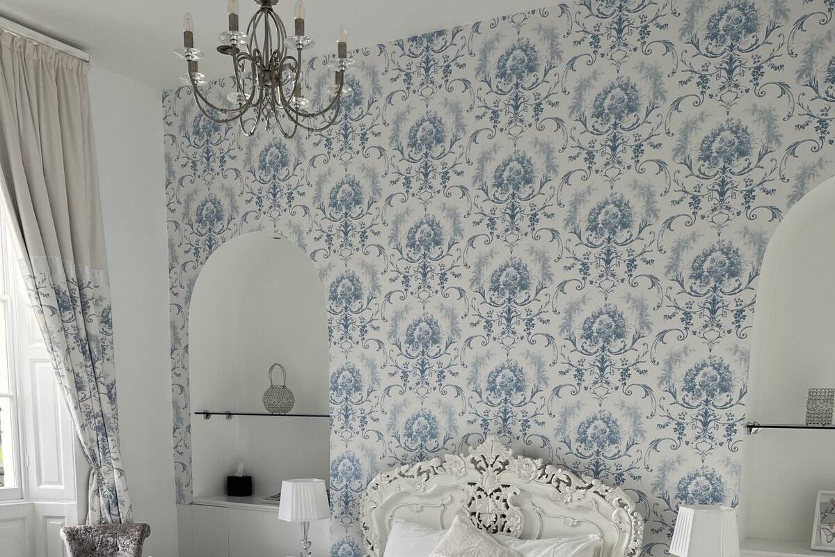 A classic bedroom with a large ornate bed, white walls, and blue floral wallpaper.