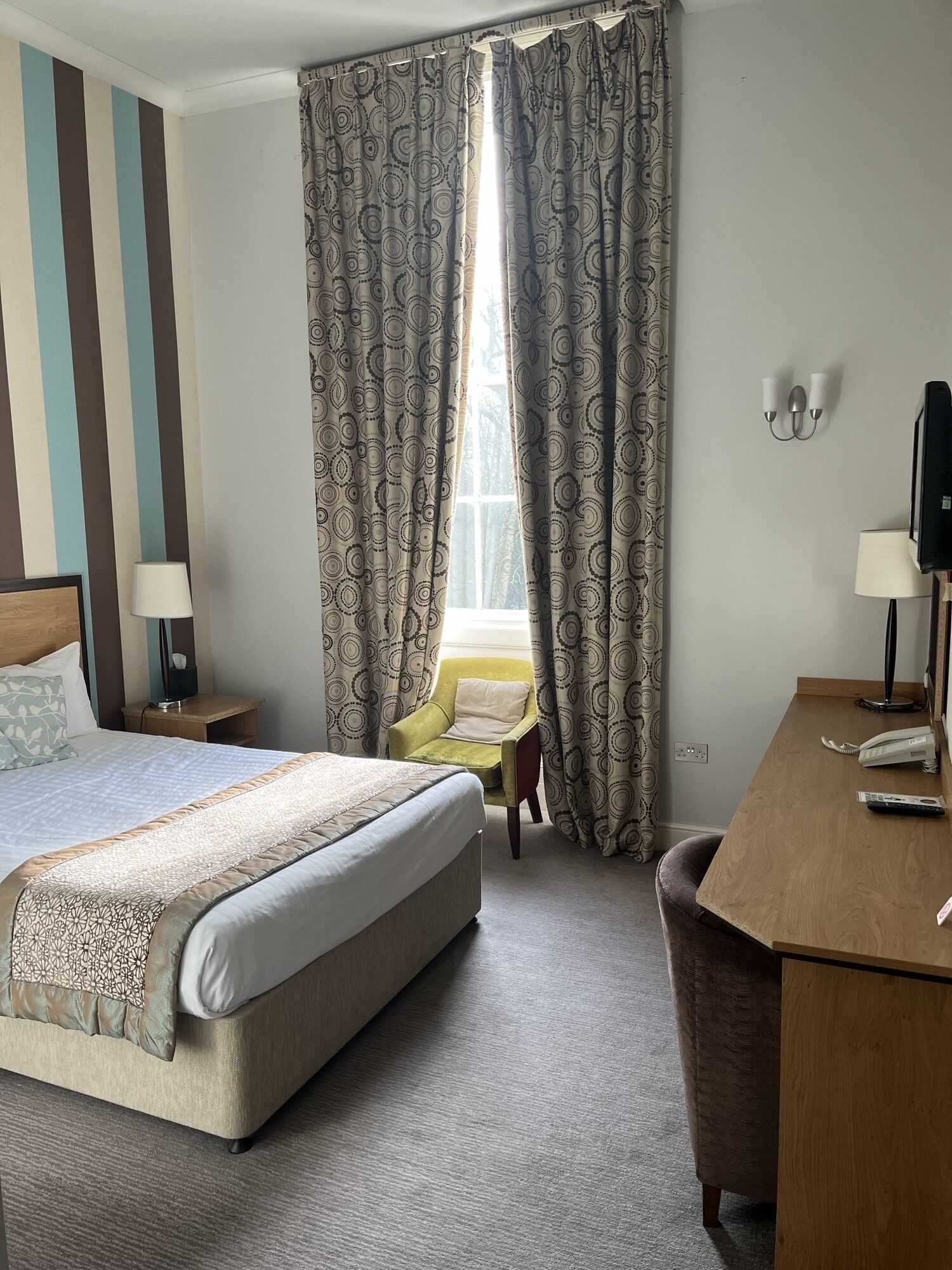 A hotel room with a large bed, a desk with a telephone, a chair, and a window with patterned curtains.