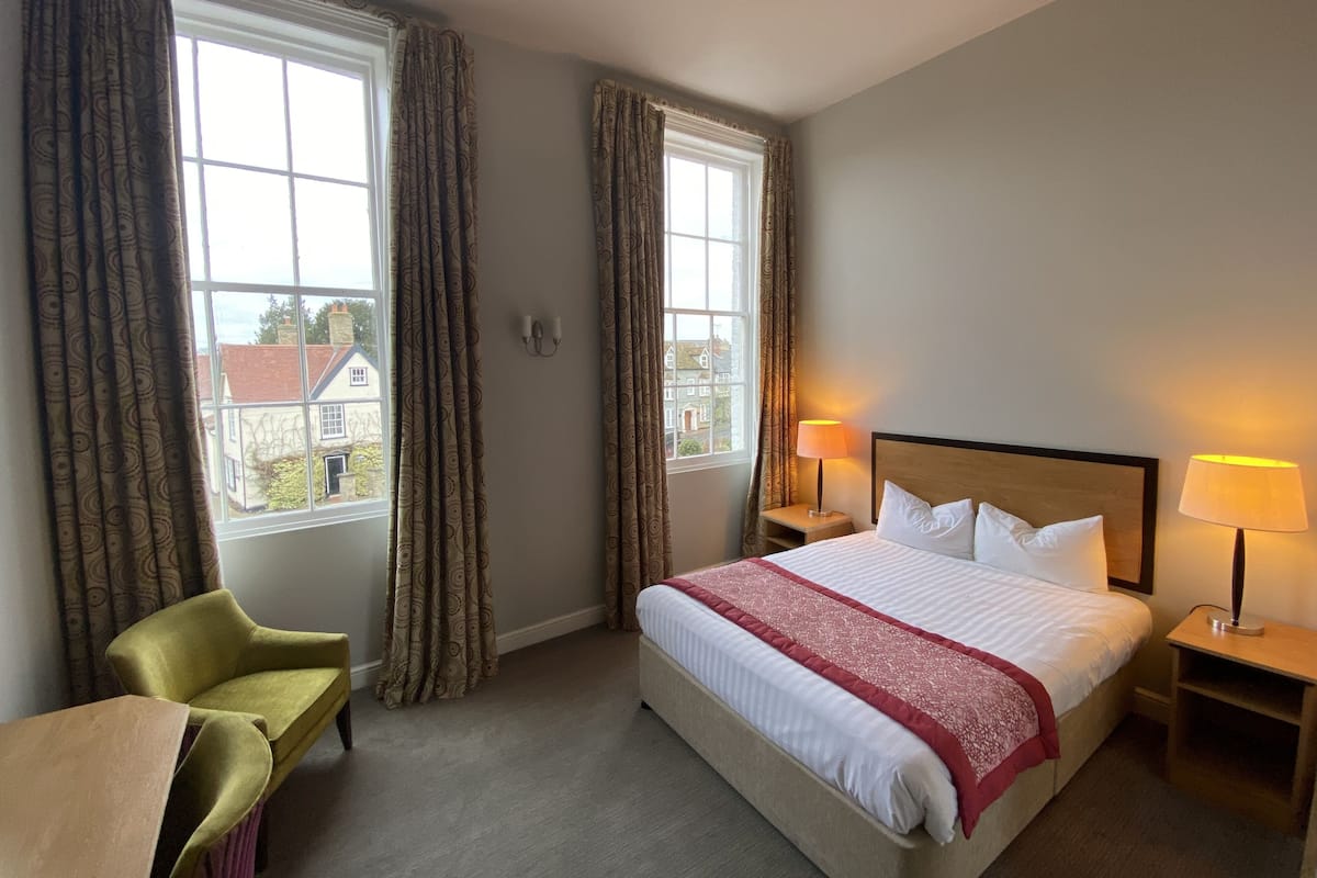 A hotel room with a bed, two bedside lamps, a chair, and two windows with curtains.