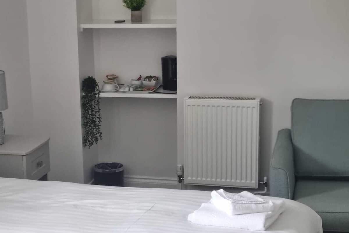 A hotel room with a bed, a nightstand with a lamp, a radiator, a chair, and a small shelf with decorative items.