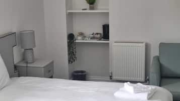 A hotel room with a bed, a nightstand with a lamp, a radiator, a chair, and a small shelf with decorative items.