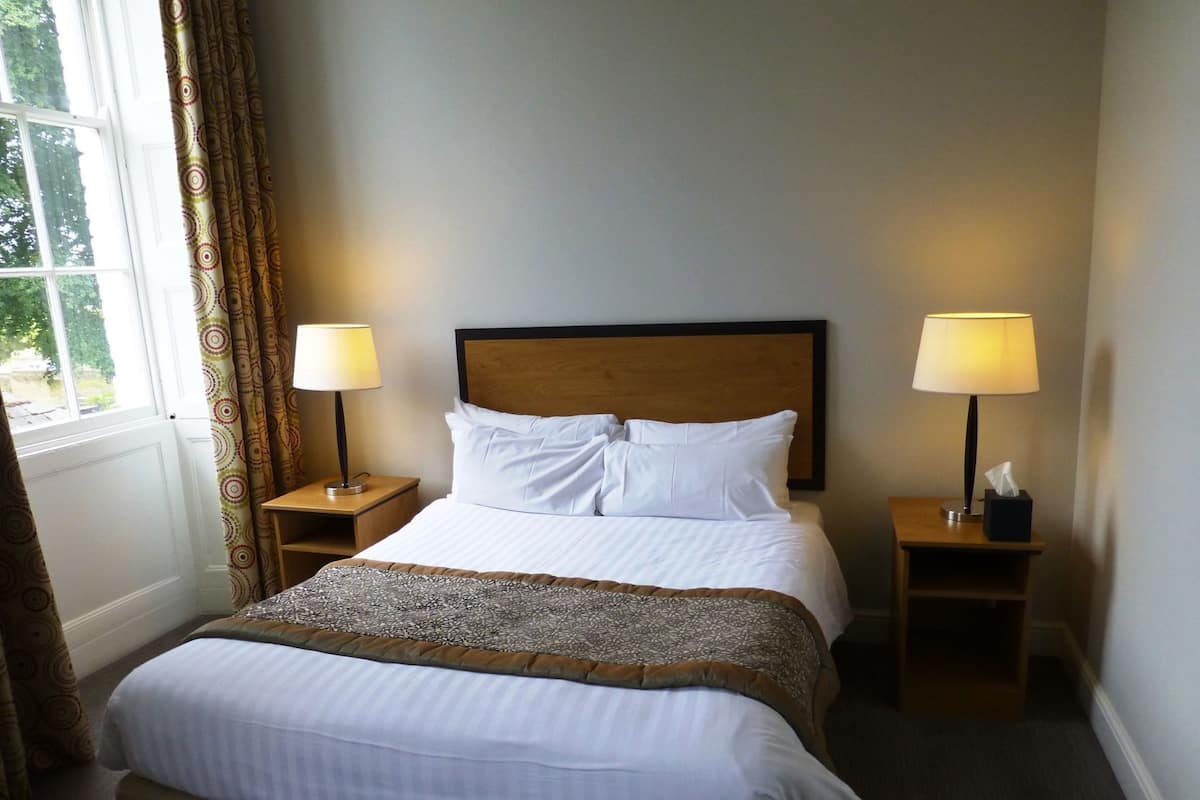 A hotel room with a bed, two bedside tables with lamps, a window with curtains, and a view of greenery outside.