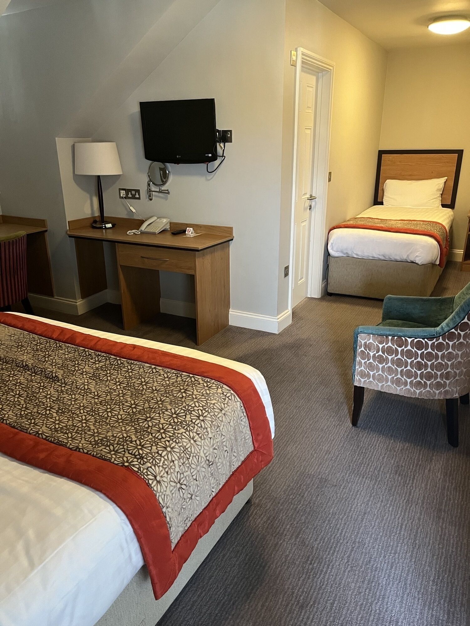 Ein Hotelzimmer mit zwei Betten, einem Schreibtisch mit Telefon, einem Sessel und einem an der Wand montierten Fernseher.