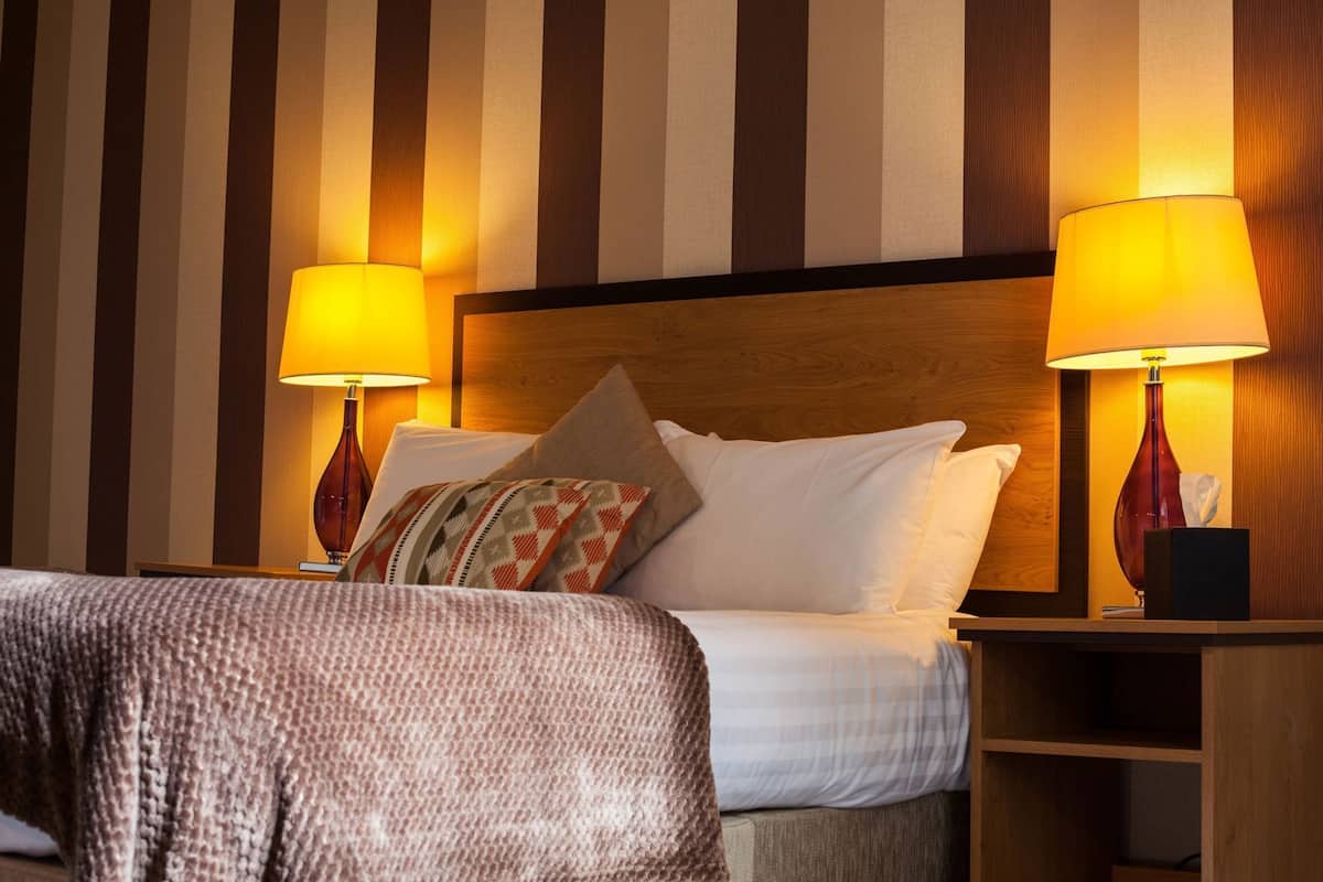 A hotel room with a bed, two bedside lamps, a wooden headboard, and striped wallpaper.