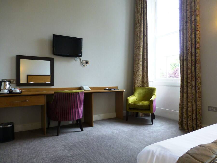 A hotel room with a wooden desk, a green chair, a bed, a TV mounted on the wall, and a window with curtains.