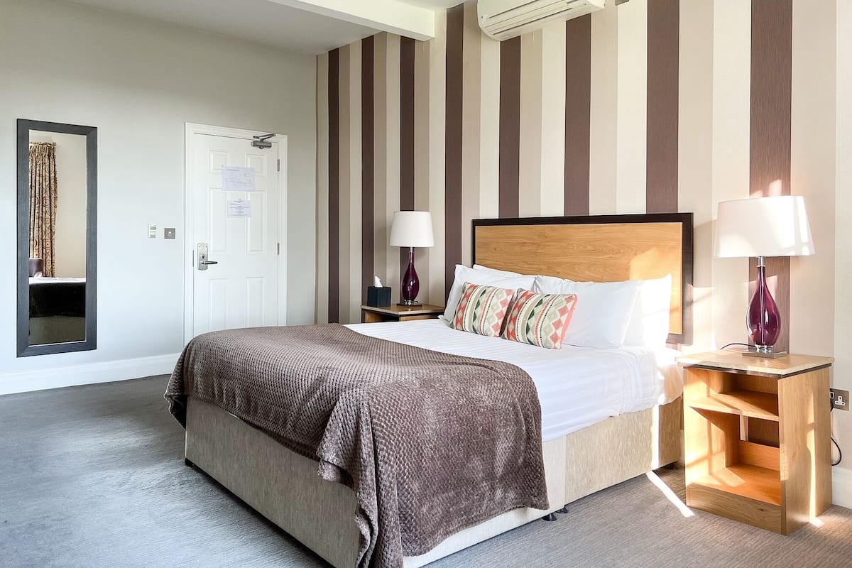 A hotel room with a bed, a nightstand, a lamp, a mirror, a door, and a wall-mounted air conditioner.